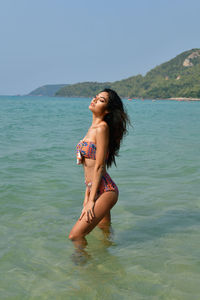 Side view of young woman wearing bikini standing in sea against clear sky