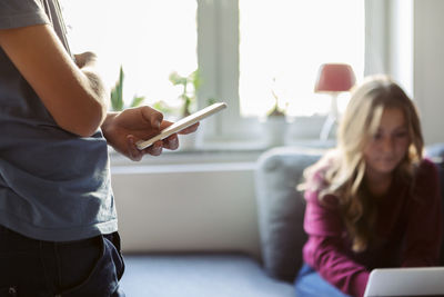 Midsection of teenager using technology while sister sitting on sofa at home