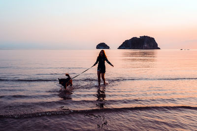 Silhouette mid adult woman with dog standing at beach against sky during sunset