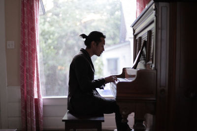 Side view of man playing piano against window at home