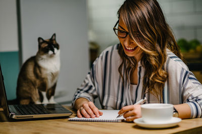 Smiling woman writing in book with cat on table 