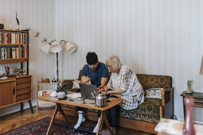 Senior woman using laptop with male care assistant sitting on sofa at home