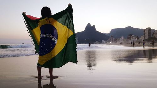 Rear view of girl holding brazilian flag while standing at beach against clear sky