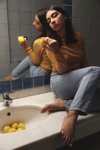 Portrait of teenage girl holding lemon while sitting against mirror on bathroom sink