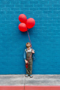 Full length of boy with red balloons standing against blue wall