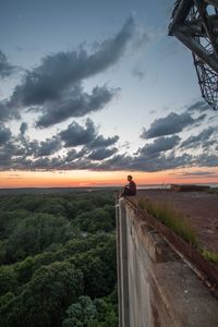 Man sitting on building terrace against sky during sunset