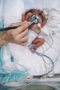 Crop doctor hand with stethoscope listening to heart anonymous newborn baby laying on hospital bed in incubator and attached to apparatus for monitoring