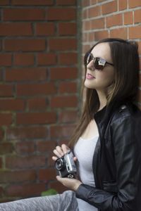 Thoughtful beautiful woman holding camera while leaning on brick wall