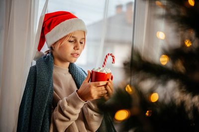 Young teen girl in santa hat having a cup of hot chocolate with marshmallow with christmas tree on