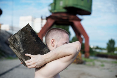 Midsection of shirtless boy holding camera against sky