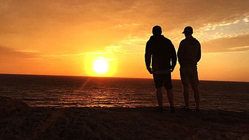 Silhouette men standing on beach against orange sky