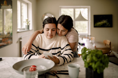 Daughter with arm around mother using smart phone at home