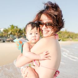 Portrait of smiling mother carrying son while standing at beach against clear sky
