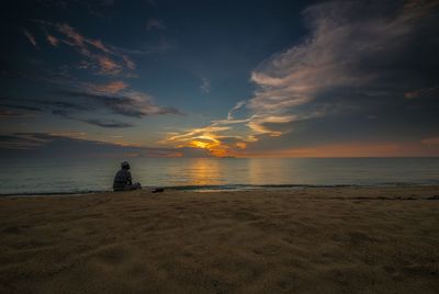 Man sitting on shore at beach against sky during sunset