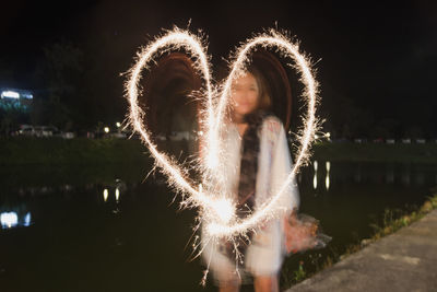 Woman making heart shape with sparkler while standing by lake against sky at night