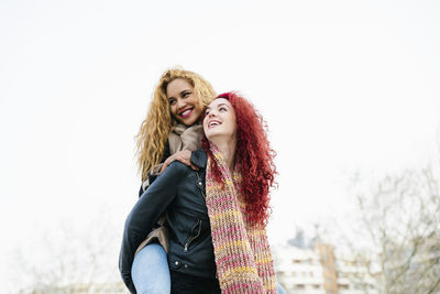 Woman piggybacking fiend while standing against clear sky