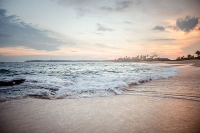 Waves rushing towards shore against sky during sunset