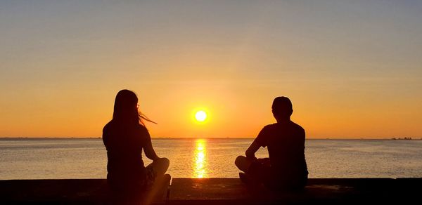 Silhouette men sitting on beach against sky during sunset