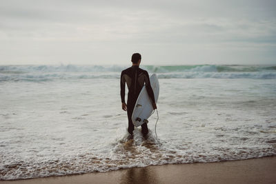 Rear view of surfer standing at beach against sky