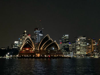 Illuminated modern buildings by river against sky at night