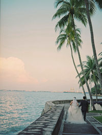 Rear view of bride and groom walking by sea against sky