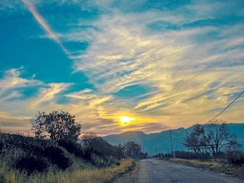 Road by trees against sky during sunset