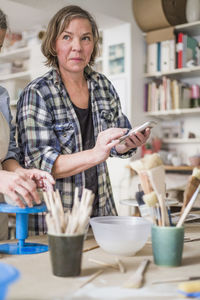 Mature female potter using mobile phone by senior colleague molding clay at workbench in store