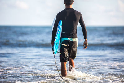 Rear view of man holding surfboard walking at beach