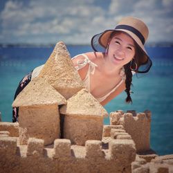Pretty asian lady behind a sand castle in a beach blue green water sea sky clouds summer smile color
