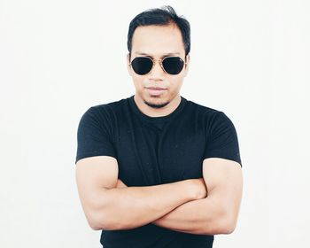 Portrait of young man with arms crossed wearing sunglasses while standing against white background