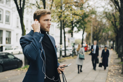 Businessman looking away while holding in-ear headphones in city