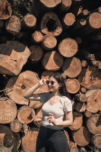 Woman holding sunglasses while standing against stack of firewood