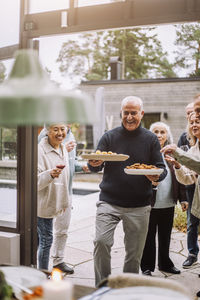 Happy mature man carrying food plates walking with friends during dinner party