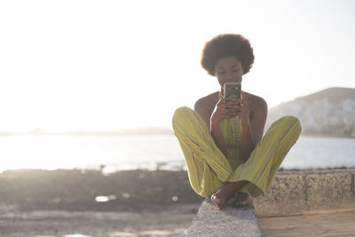 Young woman using mobile phone while sitting on retaining wall at beach against sky