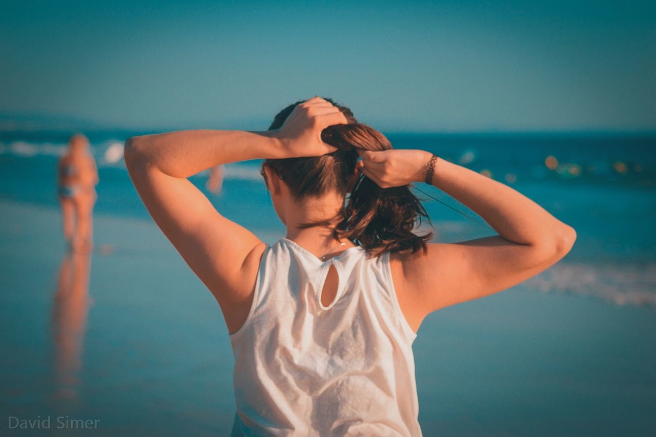 one person, real people, sea, standing, women, lifestyles, human arm, hand in hair, water, hair, adult, leisure activity, hairstyle, rear view, waist up, long hair, arms raised, casual clothing, human hair, hands behind head, beautiful woman