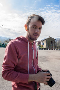 Portrait of young man holding camera while standing on street in city