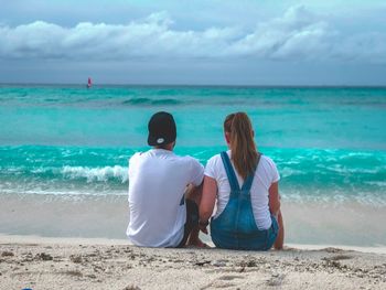 Rear view of couple sitting at beach against cloudy sky