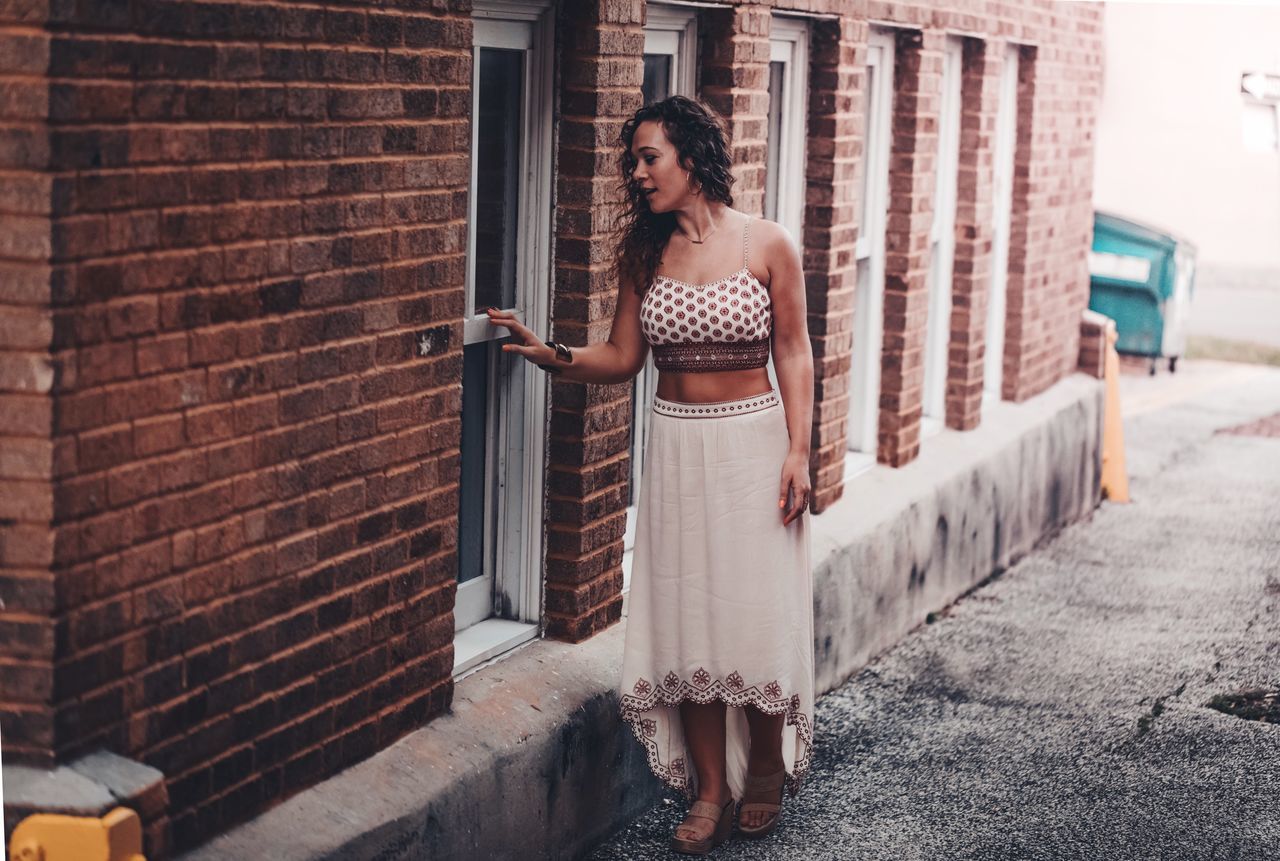 PORTRAIT OF YOUNG WOMAN STANDING BY BRICK WALL