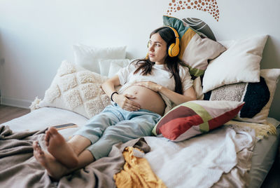 Cute brunette pregnant woman relaxes lying on the bed and listening to music on wireless headphones.