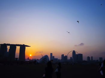 Silhouette birds flying over city against sky during sunset