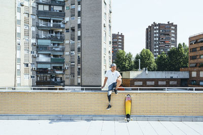 Young man with leg prosthesis sitting on railing in the city next to skateboard