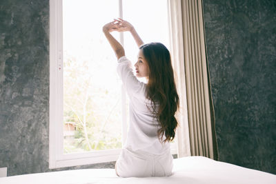 Rear view of woman stretching arms by window at home