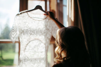 Rear view of woman holding wedding dress against window