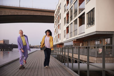 Smiling female friends or gay couple on promenade, one woman walking while other skateboarding