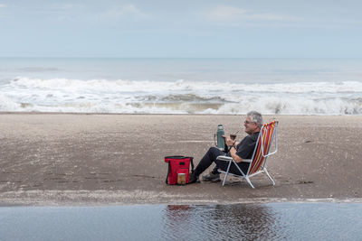 Mature man with gray hair sitting on a beach chair drinking mate and behind waves of the sea