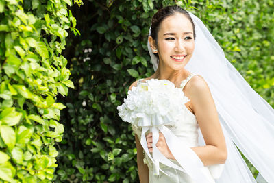 Smiling bride with bouquet standing against plants 