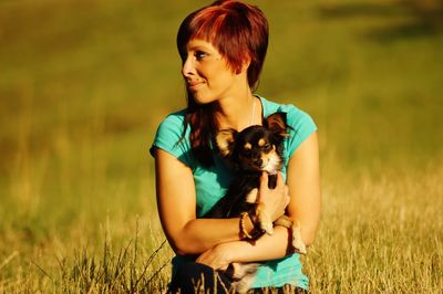 Smiling redhead woman carrying dog while sitting on grassy field