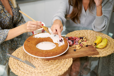 Crop anonymous female friends garnishing baked vegan banana cake with topping and fresh berries and nuts in home kitchen