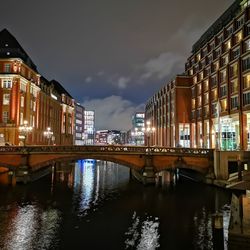 Bridge over river by illuminated buildings against sky at night