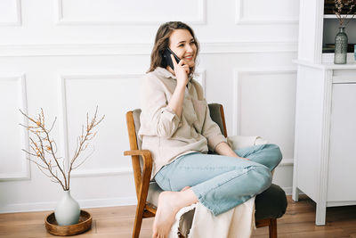 Millennial woman smiling, speaking on phone, sitting on chair, looking out window at free space